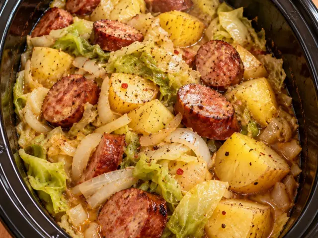 Slow cooker filled with smoked sausage, cabbage, potatoes, and onions in broth