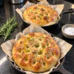 Two golden mini rosemary olive oil focaccias in parchment-lined cast-iron skillets