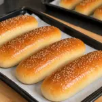 Golden sesame hot dog buns on a black parchment-lined baking tray